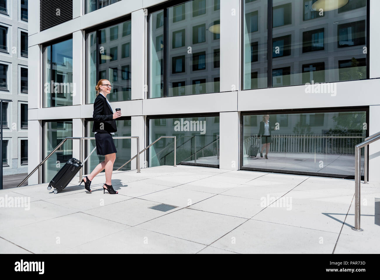 Imprenditrice sorridente con la valigia e caffè per andare a piedi nella parte anteriore dell'edificio degli uffici Foto Stock