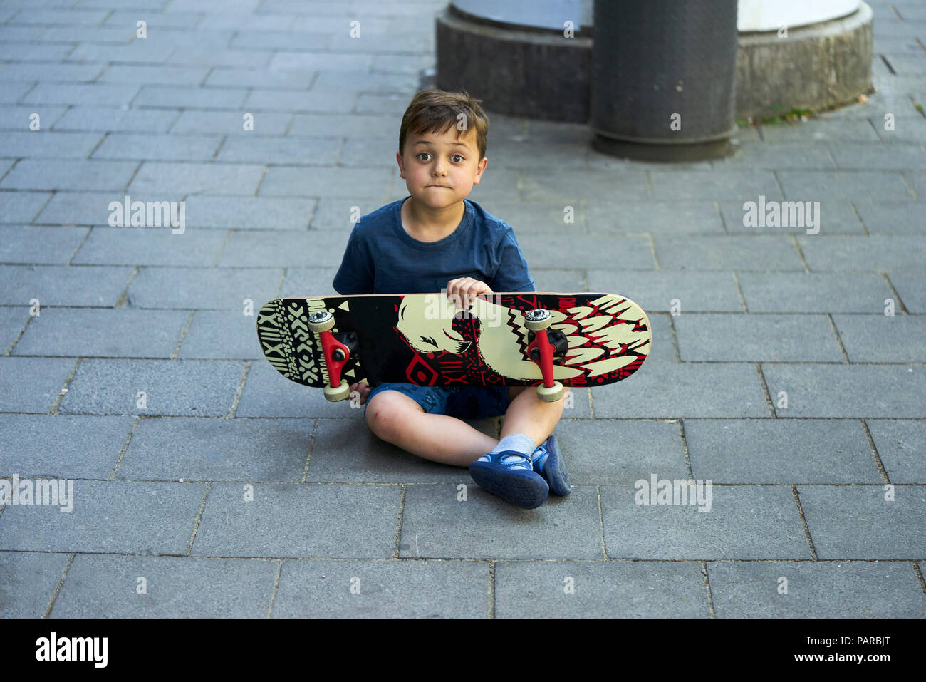 Ritratto di stupito piccolo ragazzo seduto sul marciapiede con lo skateboard Foto Stock