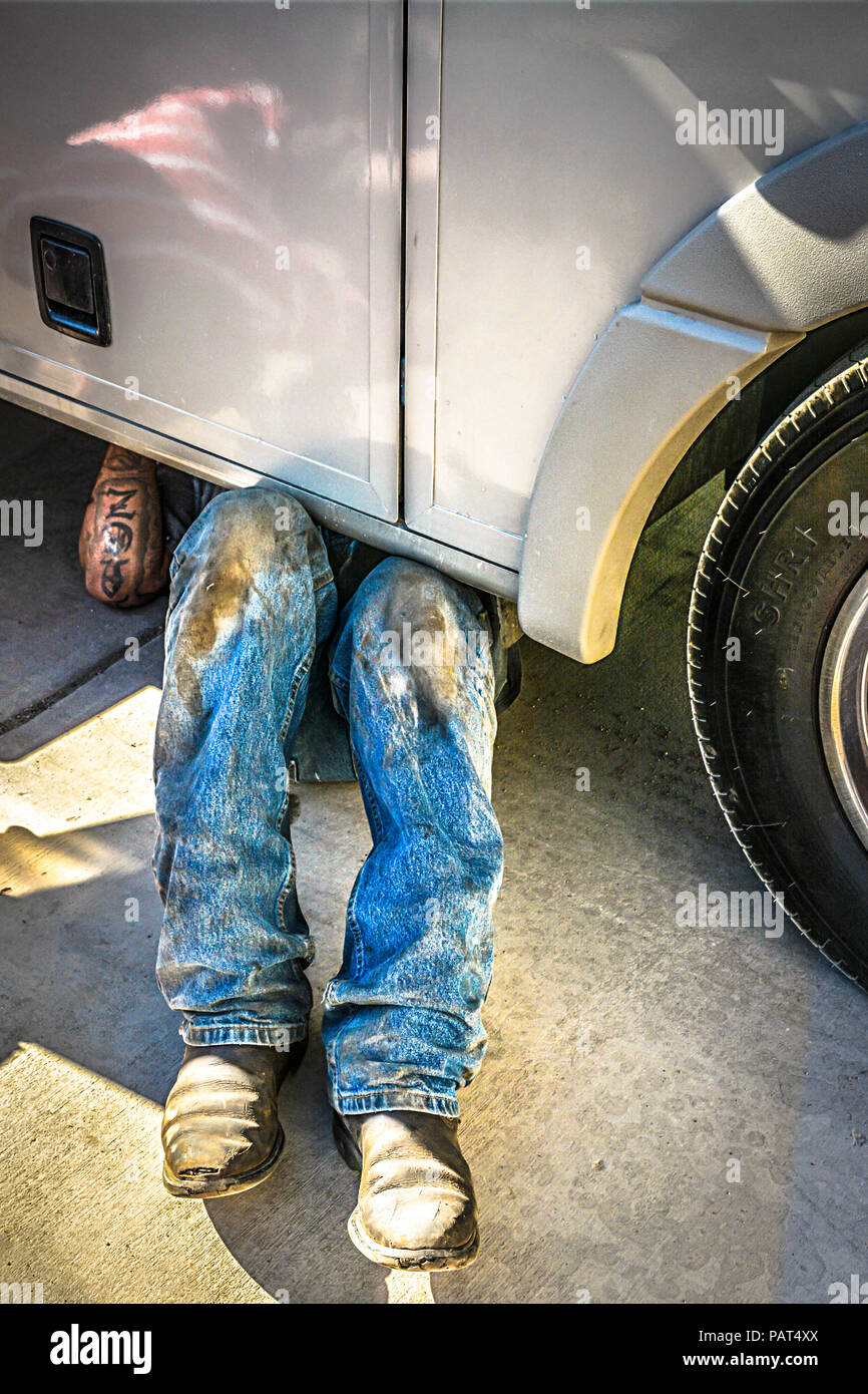 Un meccanico sotto il carrello indossare stivali da cowboy e sporca jeans visto solo dalla vita scende al di fuori servizio garage in Nuovo Messico Foto Stock