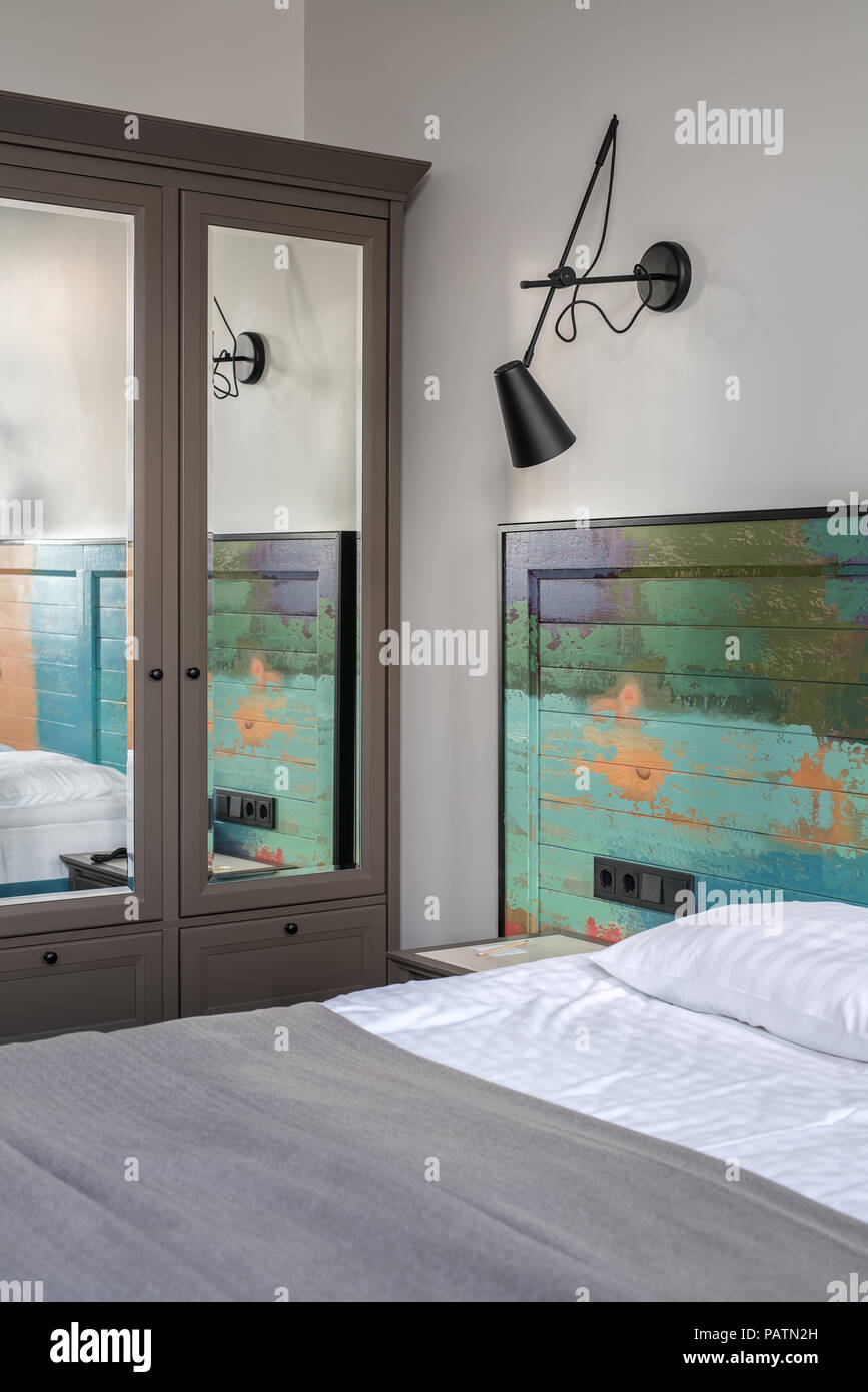 Bella camera in un albergo con pareti chiare. Vi è un letto con un coloratissimo testata in legno e lenzuola bianche con un coverlet sul letto, comodino, nero lampada Foto Stock