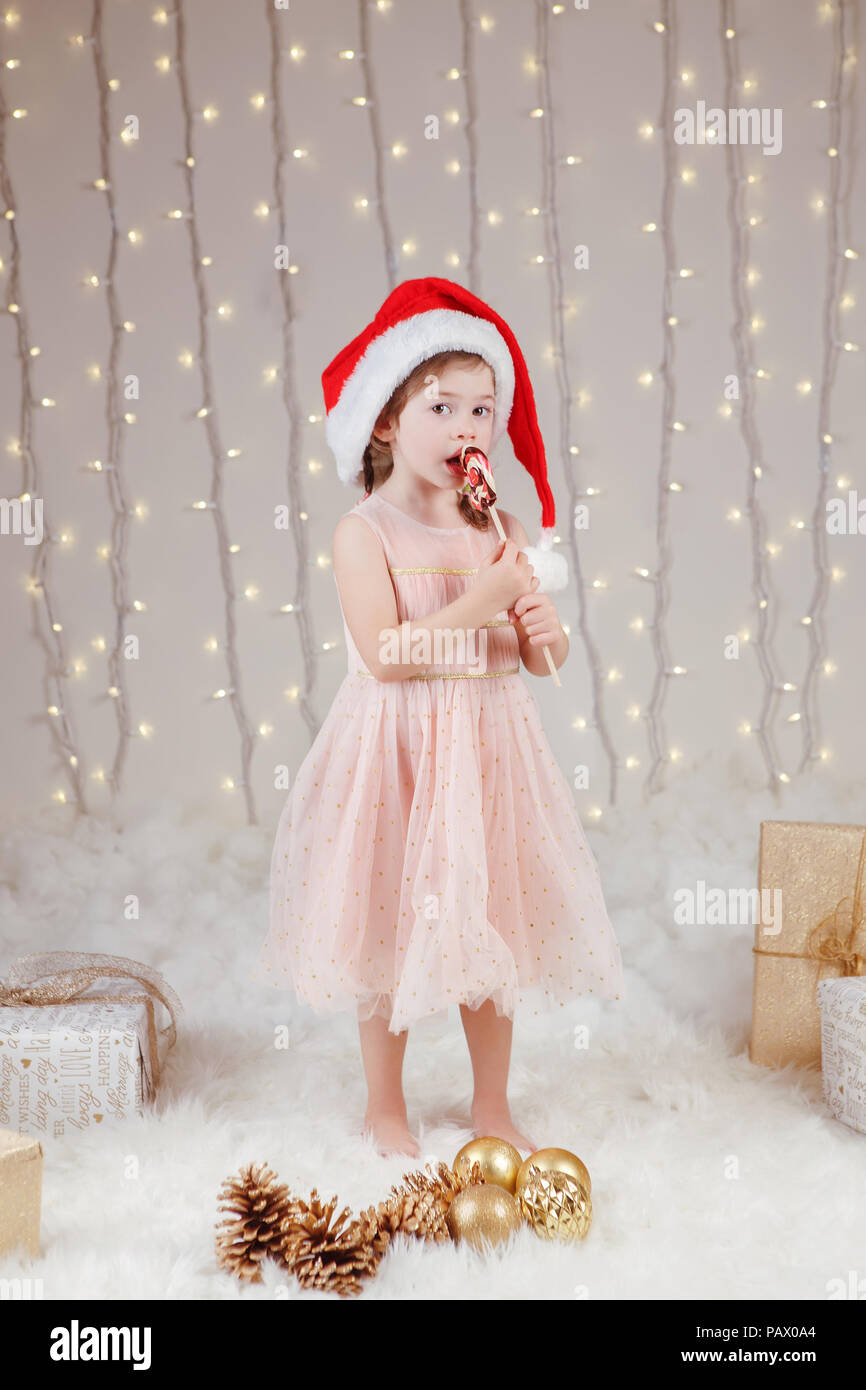 Ritratto di bianco ragazza caucasica indossando Santa Claus hat celebrando il Natale o il nuovo anno. Poco carino adorabile bambino mangiare leccare la caramella lecca-lecca i Foto Stock