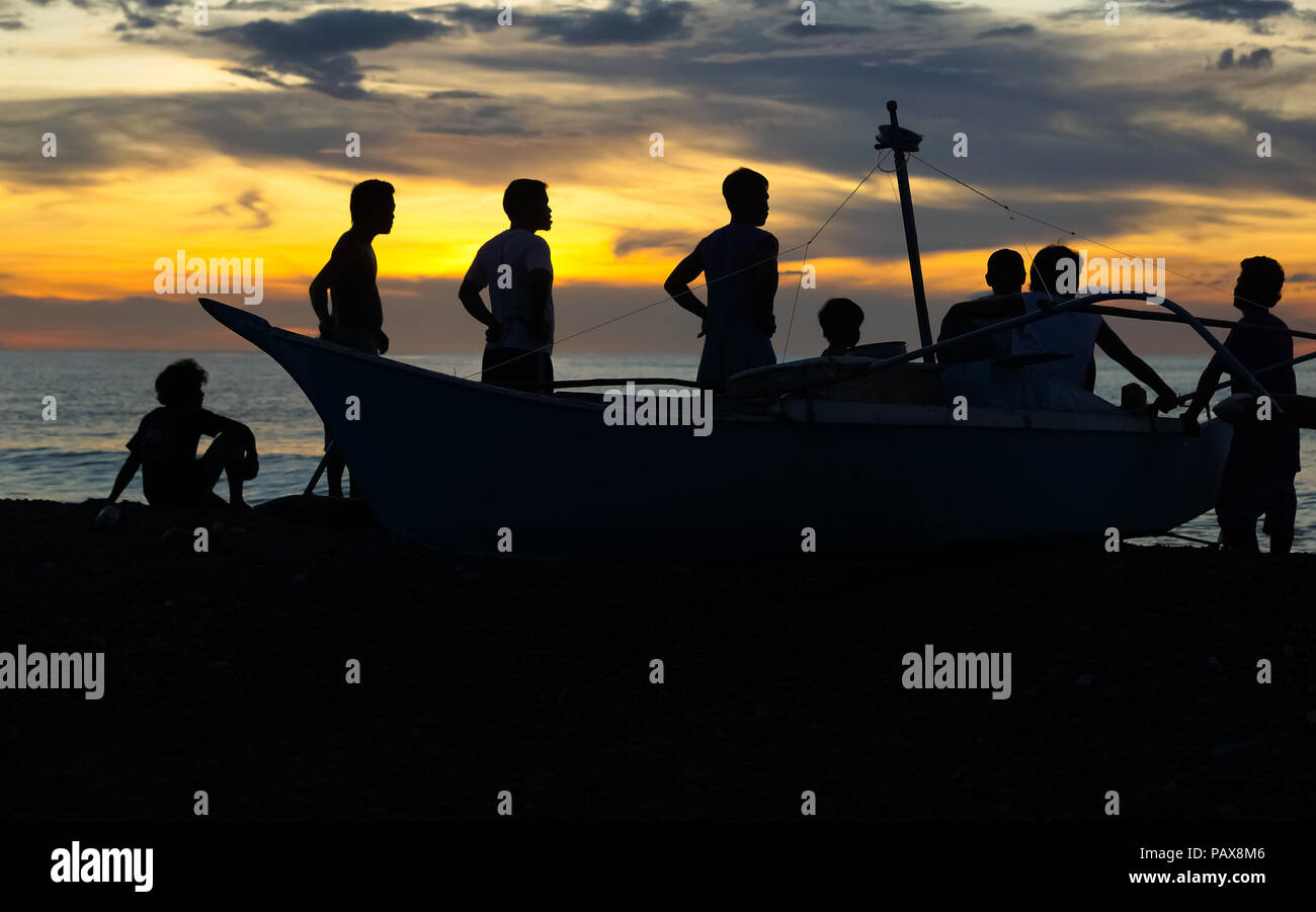 In piedi e seduta gruppo di pescatori con barca da pesca silhouette isola sulla spiaggia al tramonto - Panay, Filippine Foto Stock