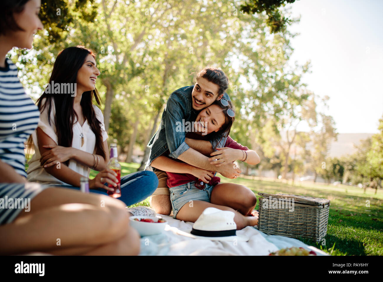 Uomo che abbraccia la sua ragazza mentre è seduto con amici di sesso femminile al parco. Felice giovani amici gustando un picnic. Foto Stock
