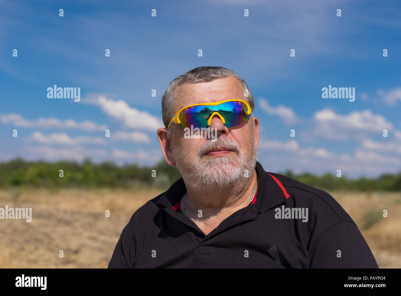 Outdoor ritratto di un barbuto senior in occhiali da sole contro blu cielo nuvoloso e il settore agricolo. L'uomo è in cerca di speranza Foto Stock