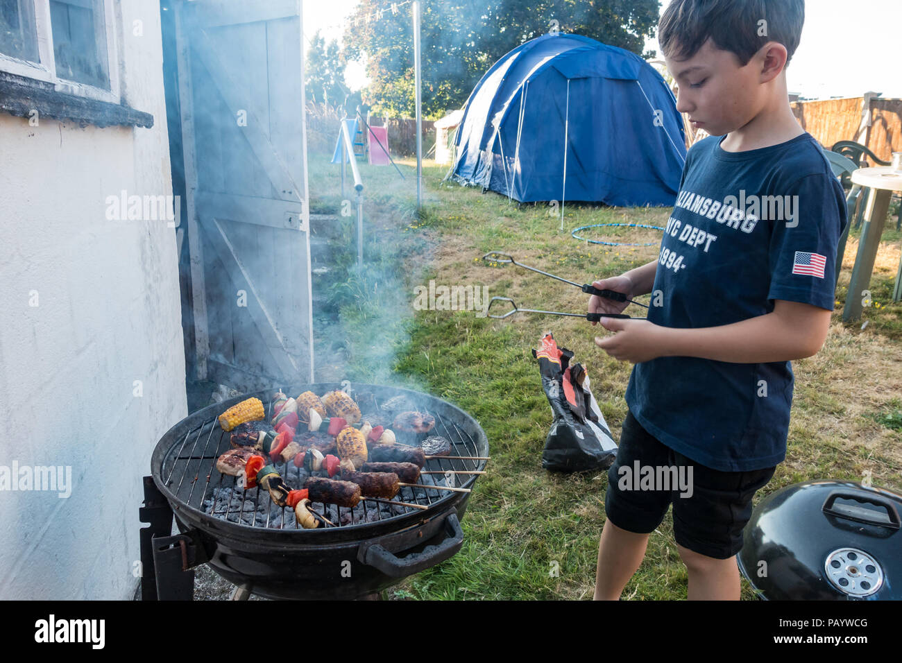 Un giovane ragazzo frequenta un barbecue in giardino sul retro su cui gli hamburger, spiedini e verdure sono in cottura. Foto Stock