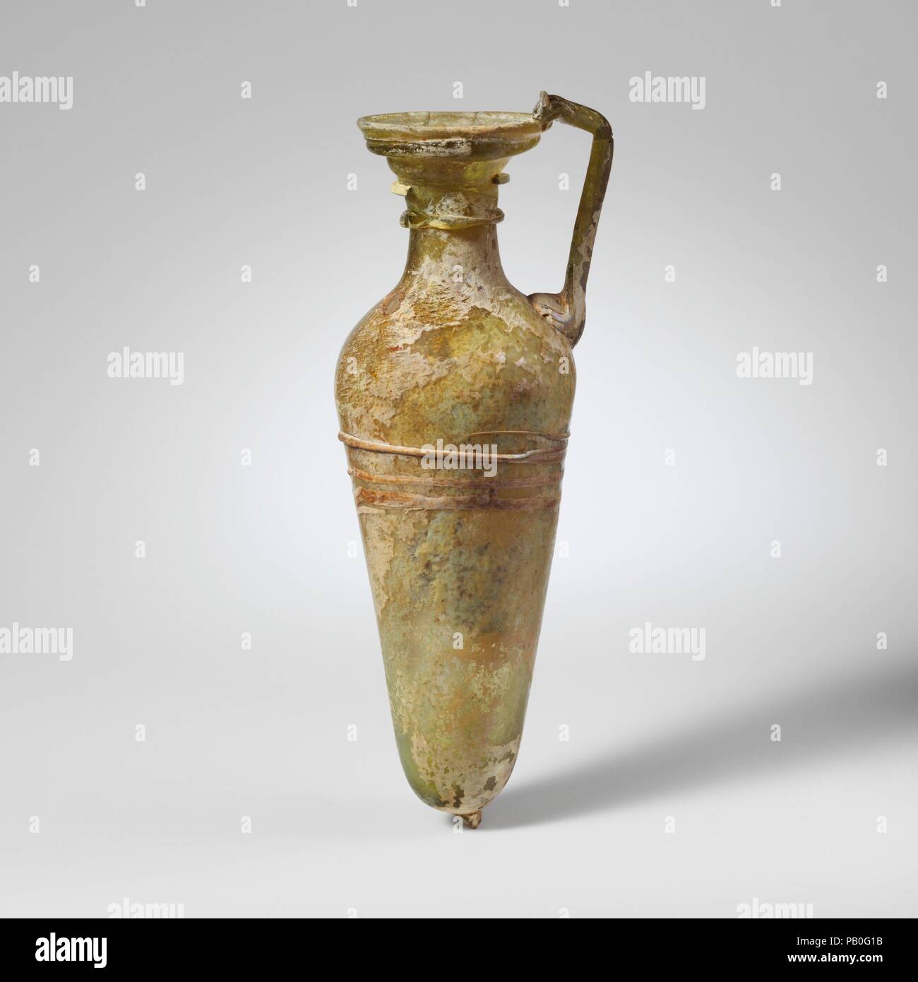 Caraffa di vetro. Cultura: Romano. Dimensioni: complessivo: 7 7/16a. (18.9cm). Data: 4th-5secolo D.C.. Traslucido giallo verde; la maniglia e sentieri nello stesso colore ma una scia di colore incerto, possibilmente bianco opaco. Cerchione piegato sopra e in; ovale, bocca svasata, con attrezzature trattini; collo ovale, si espande verso il basso; spalla spiovente; alto e snello corpo con lato rettilineo con la conicità rivolta verso il basso; fondo arrotondato con prominenti pontil tampone; impugnatura asta fissata come un tampone di grandi dimensioni a spalla, redatto e leggermente fuori e poi trasformato in e fatta cadere sulla parte superiore del cerchio. Un sentiero avvolto in senso orizzontale una volta e mezza un Foto Stock