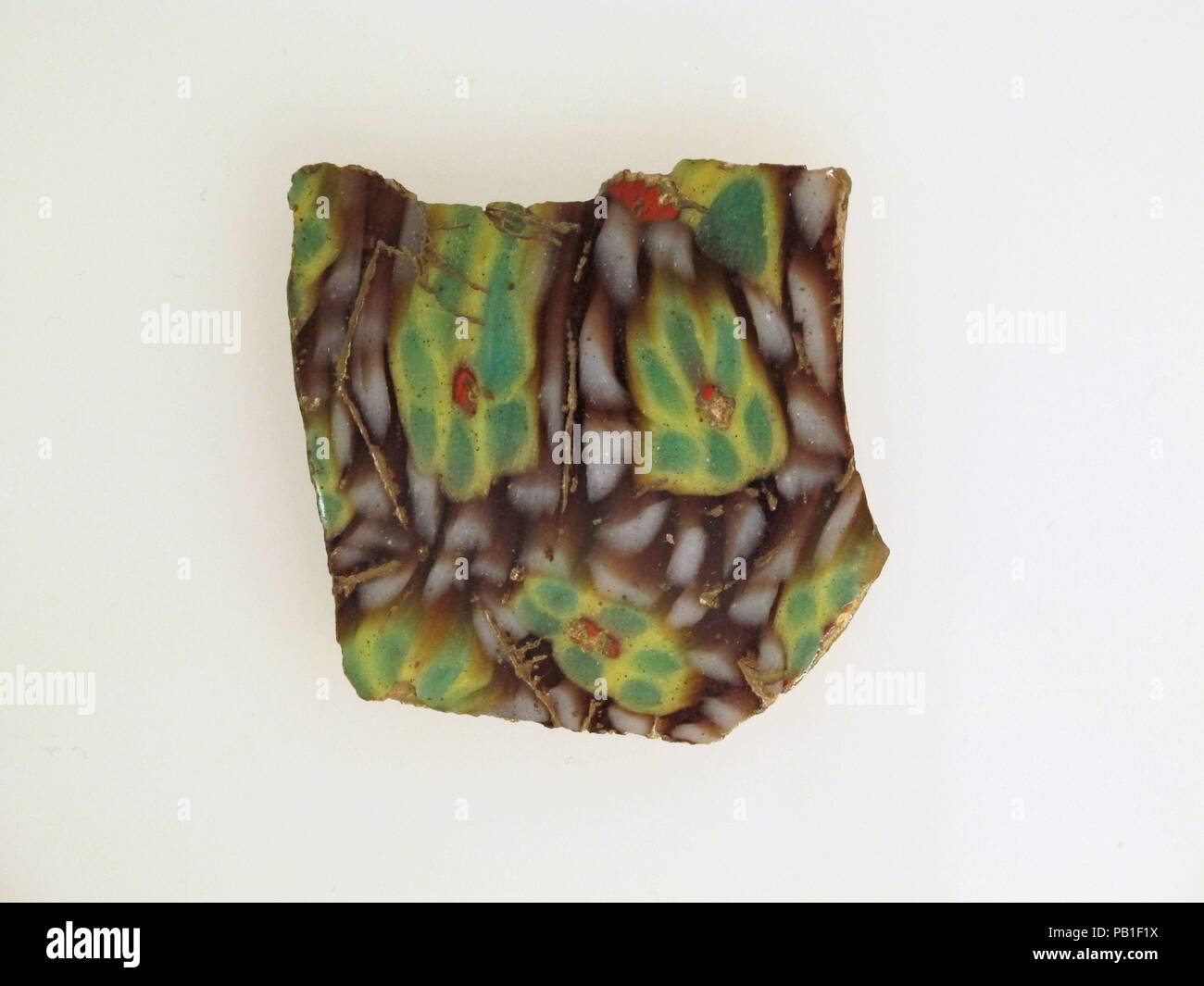 Mosaico di vetro frammento. Cultura: Romano. Dimensioni: 1 3/4 × 1 11/16 × 1/8 in. (4,5 × 4,3 × 0,3 cm). Data: fine del I secolo A.C. - inizio del I secolo d.c. Traslucidi viola, giallo opaco, bianco, rosso e verde. La curvatura convessa lato, eventualmente con parte di una verticale, bordo smussato da un grande piatto. Mosaico formata da grandi sezioni poligonali di una singola canna da zucchero in una massa di colore viola con un cerchio di aste bianche che circondano la configurazione a rosetta delineata in giallo comprendente sei petali di colore verde e uno rosso centrale asta. Lucido esterno; lievi tracce di vaiolatura e agli agenti atmosferici di bolle di superficie e si unisce tra Foto Stock
