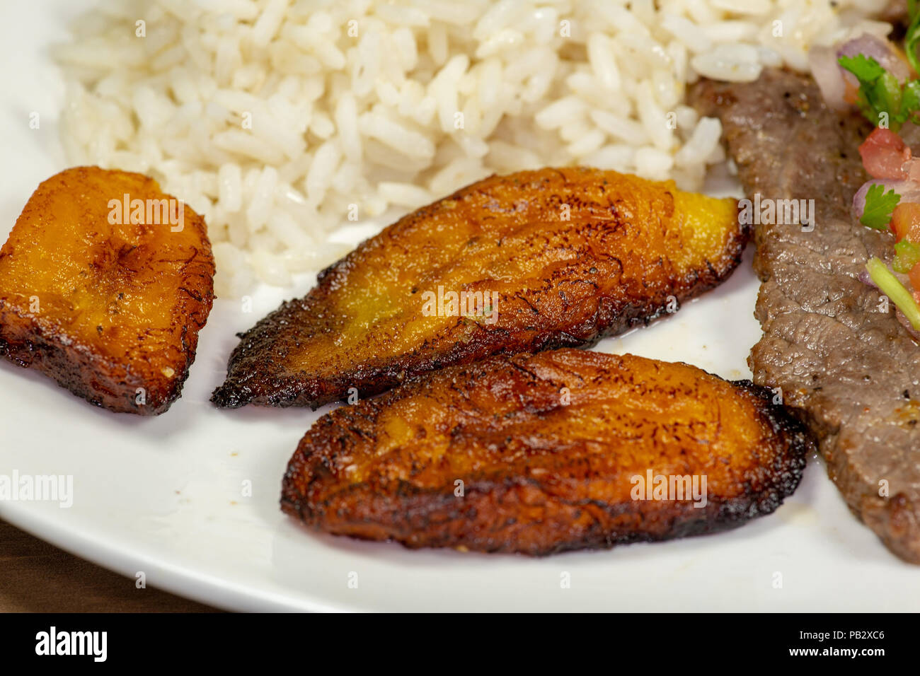 Una bistecca coperto di Pico de Gallo circondato da platani e riso bianco su una piastra bianca. Cibo cubano. Foto Stock