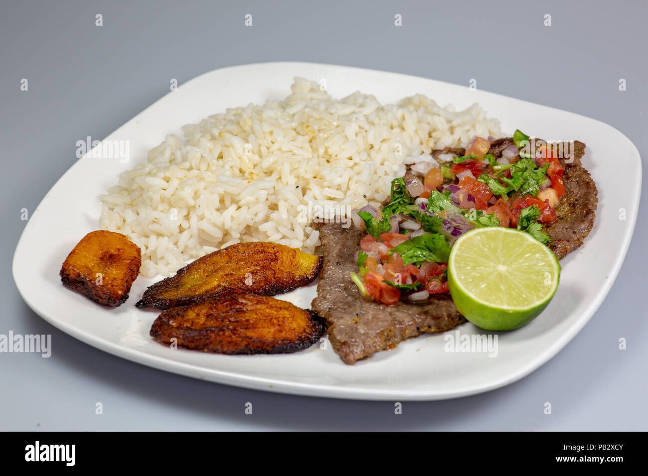 Una bistecca coperto di Pico de Gallo circondato da platani e riso bianco su una piastra bianca. Cibo cubano. Foto Stock