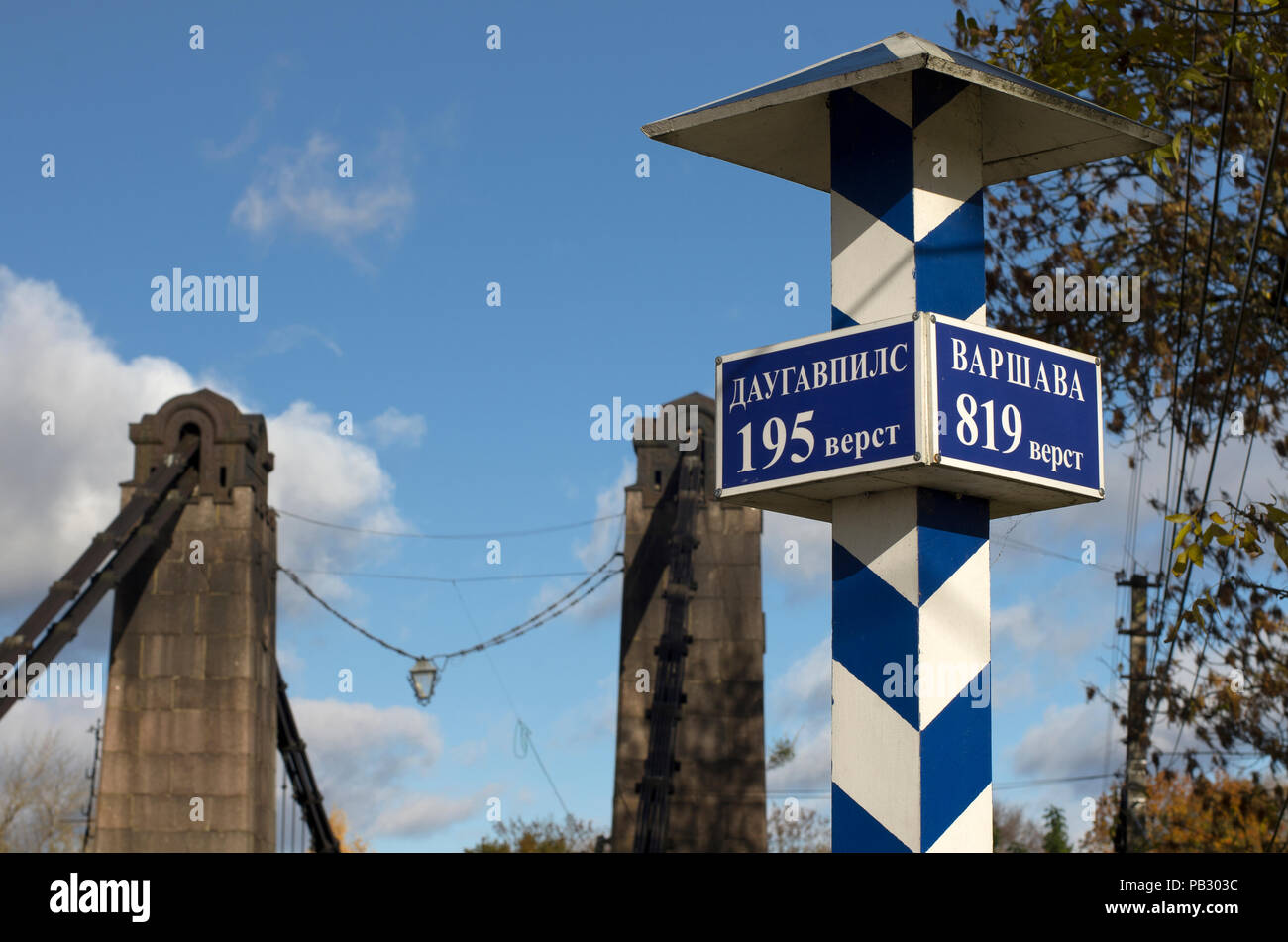 Milepost con i nomi delle città in russo e la distanza in chilometri e i pilastri del ponte di sospensione in background Foto Stock