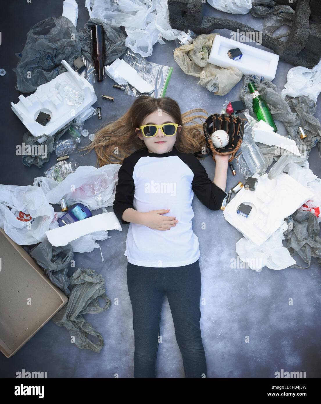 Un giovane bambino sta tenendo in mano una palla da baseball con rifiuti Rifiuti come sacchetti di plastica e non di prodotti biodegradabili intorno a lei per un riciclo energia pulita concept Foto Stock