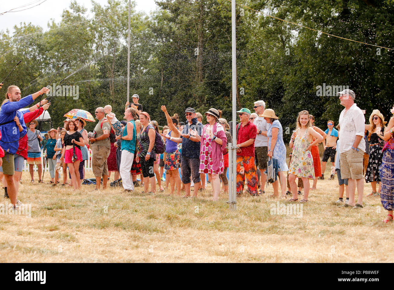 Charlton Park di Malmesbury, Wiltshire. Il 27 luglio 2018. Folle sono sorpresi da un teatro di strada delle prestazioni mediante il gruppo Commandeau Compagnie Pernette. Gli interpreti utilizzano acqua giardino irroratrici nella danza sincronizzata di Stauss il Danubio blu - mentre spruzzando la folla con acqua nel processo! Molto apprezzato dal pubblico in condizioni di caldo. Il festival prende il via in condizioni calde e umide per la sua prima giornata piena di musica del mondo, il teatro di strada e workshop. Credito: Wayne Farrell/Alamy Live News Foto Stock