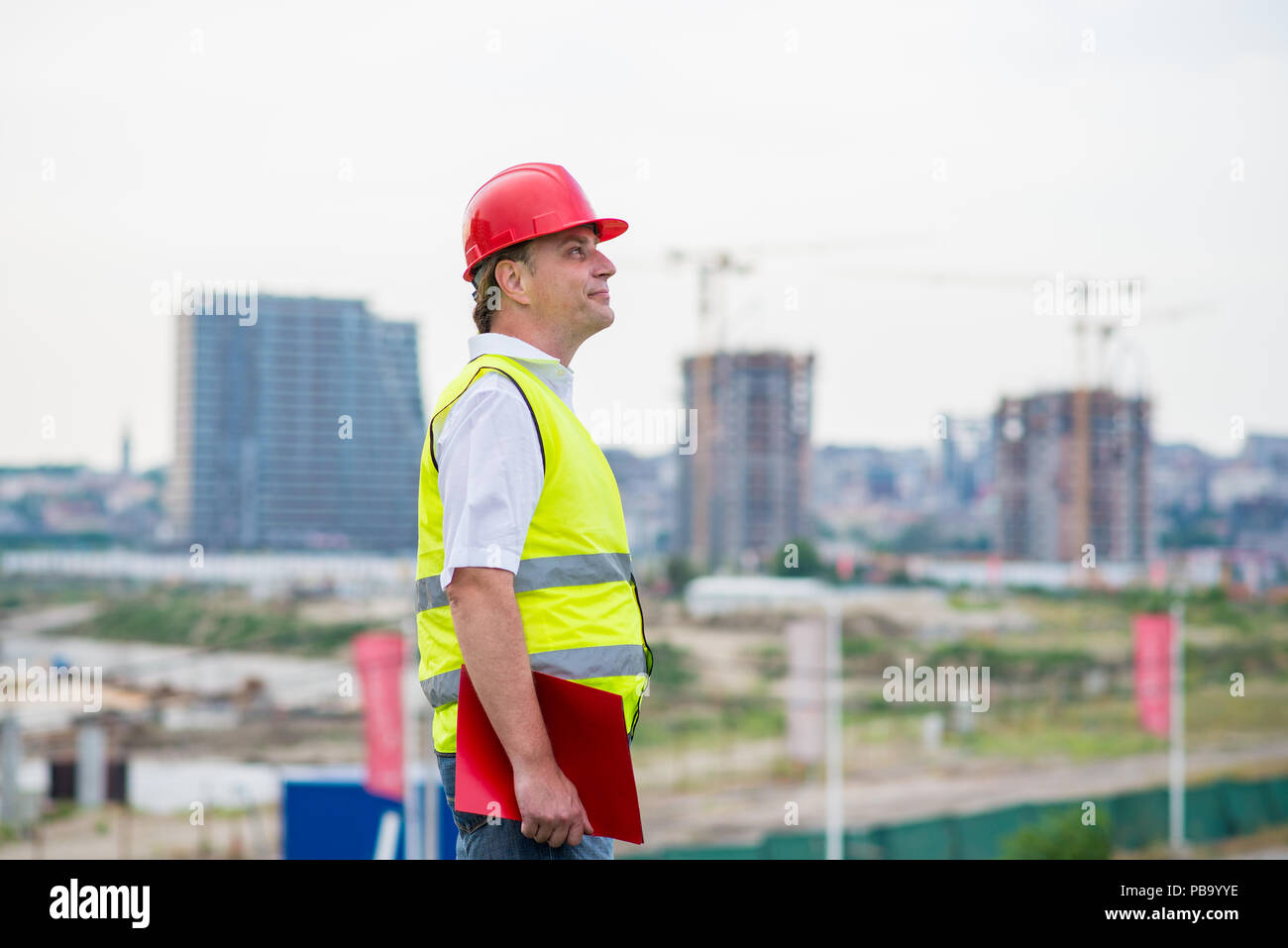 Tecnico su un sito in costruzione che indossa giallo riflettente west e casco rosso con edifici e gru in background. Caposquadra su un sito di costruzione Foto Stock