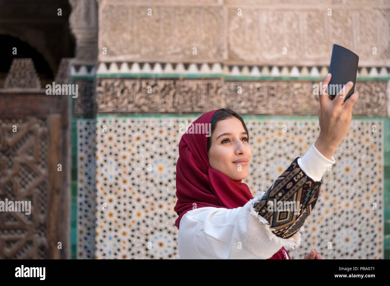 Giovane donna musulmana tenendo selfie con telefono cellulare in abbigliamento tradizionale con velo di colore rosso Foto Stock