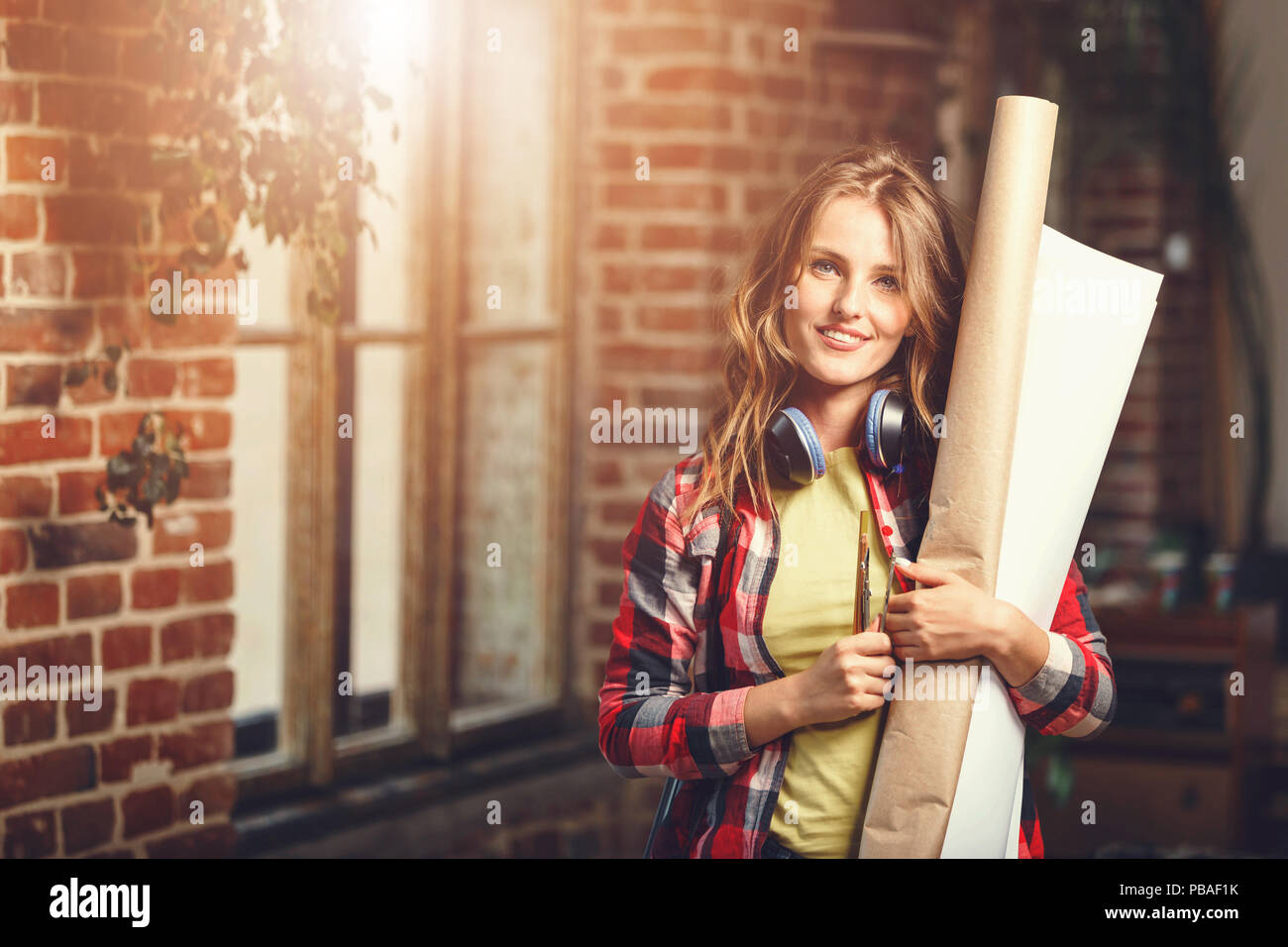 Bel giovane a lungo i capelli architetto femmina holding rotoli di carta e sorridenti vicino alla finestra in mansarda ufficio moderno Foto Stock