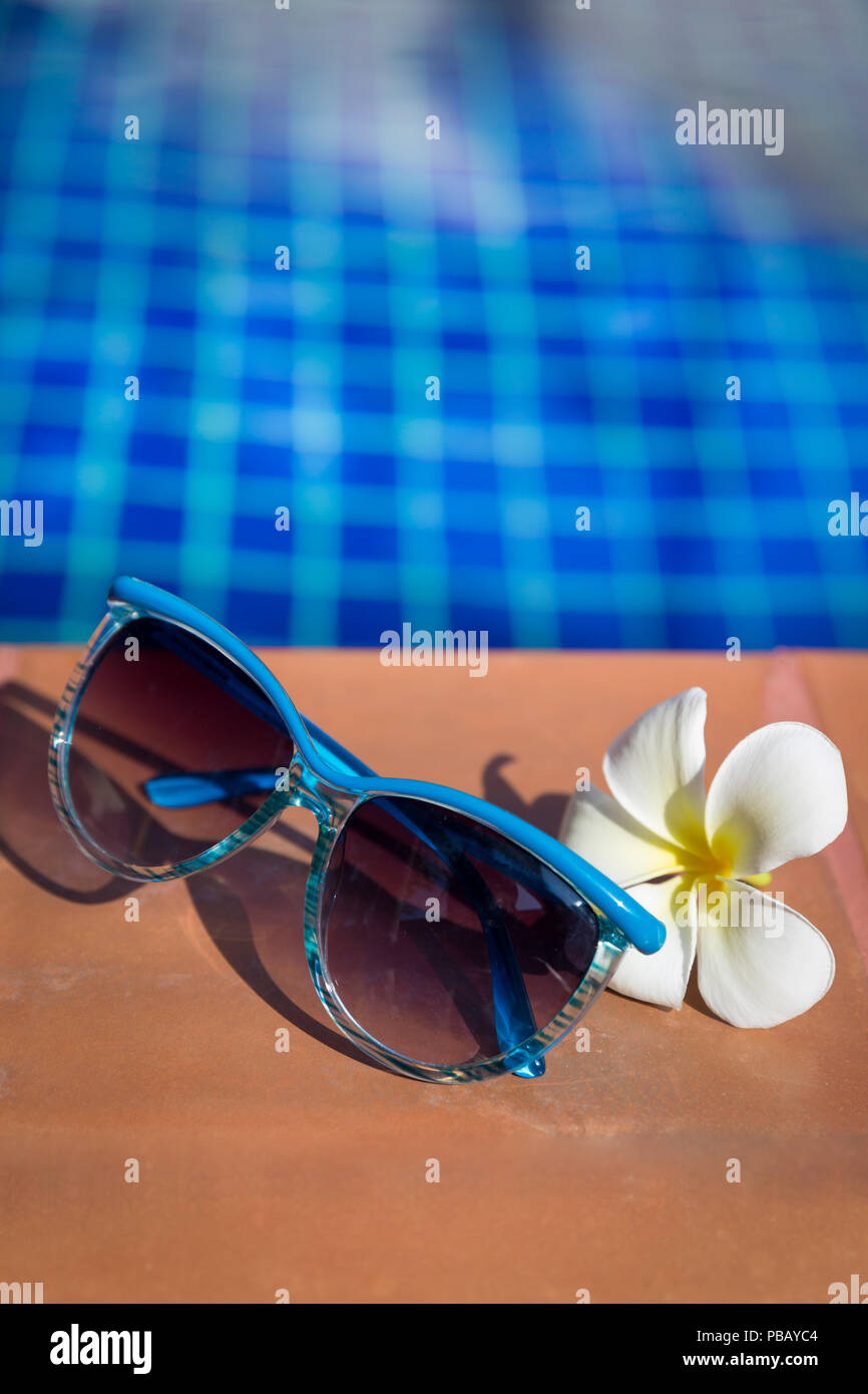Occhiali da sole blu con la plumeria, frangipani fiore sul bordo di una piscina - vacanza concetto tropicale Foto Stock