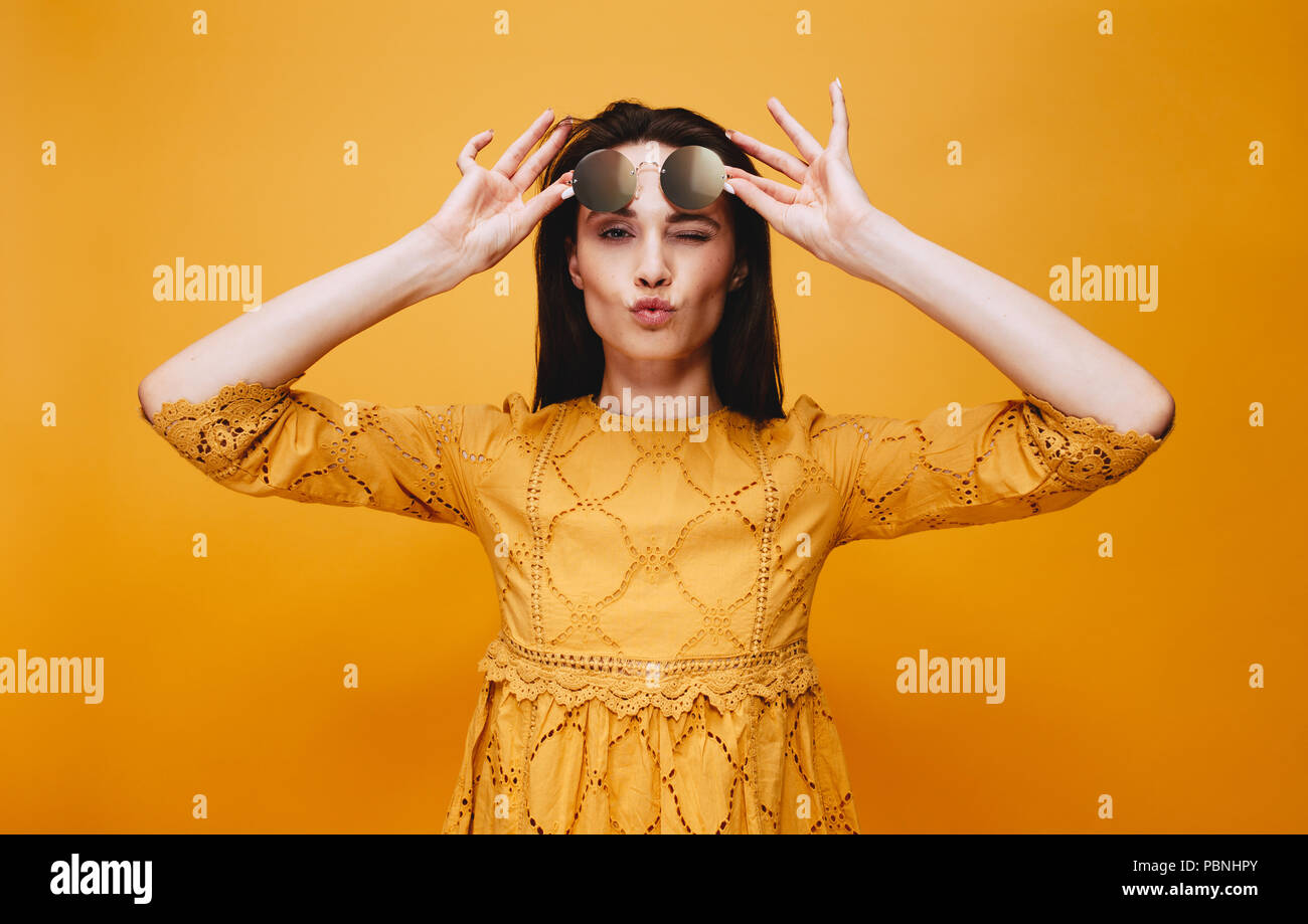Ritratto di donna che indossa abiti di colore arancione tenendo gli occhiali da sole sul fronte, winking e soffiando un bacio. Moda modello femminile in posa con labbra rugose Foto Stock