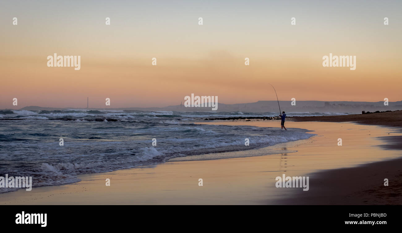 Un panorama al tramonto di una silhouette di un pescatore distante la pesca al largo di una deserta spiaggia australiana, australia Foto Stock
