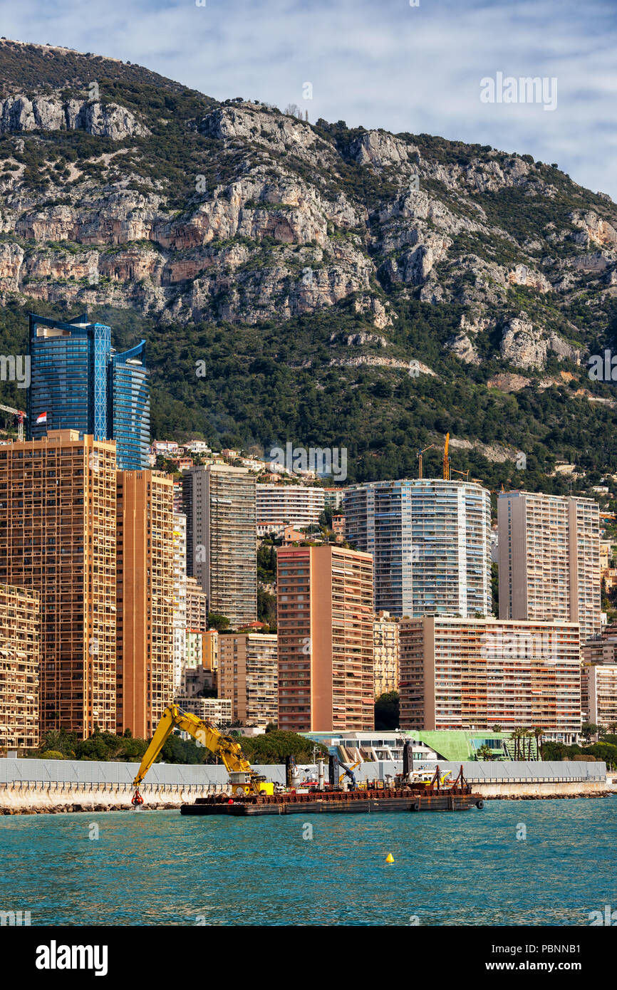 Principato di Monaco dal mare, chiatta con gru rafforza la diga frangionde, appartamento torri, i blocchi di appartamenti contro una montagna Foto Stock