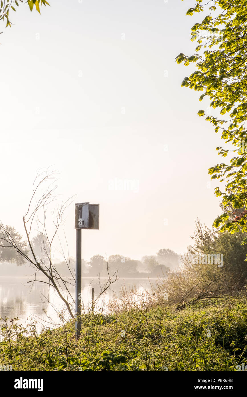 Alba riverside scena. Alberi e un chilometro di distanza marcatore sul lungofiume in una nebbiosa mattina di primavera sul fiume Trent, Nottinghamshire, England, Regno Unito Foto Stock
