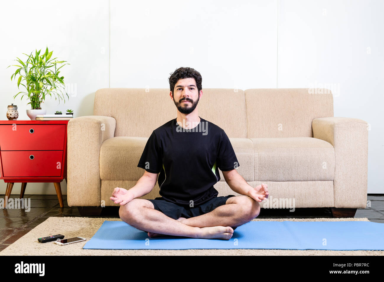 Bella caucasica uomo barbuto vestiti di nero su blu yogamat facendo siddha postura con gli occhi aperti Foto Stock