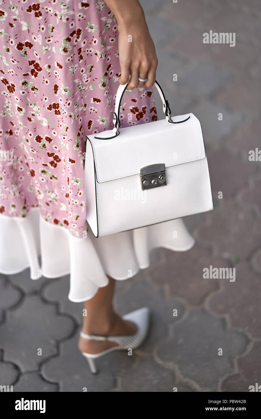 La ragazza è in posa con una borsa bianca su sfondo di strada all'aperto. Indossa un abito rosa con stampe floreali e scarpe di luce. Closeup foto verticale. Foto Stock