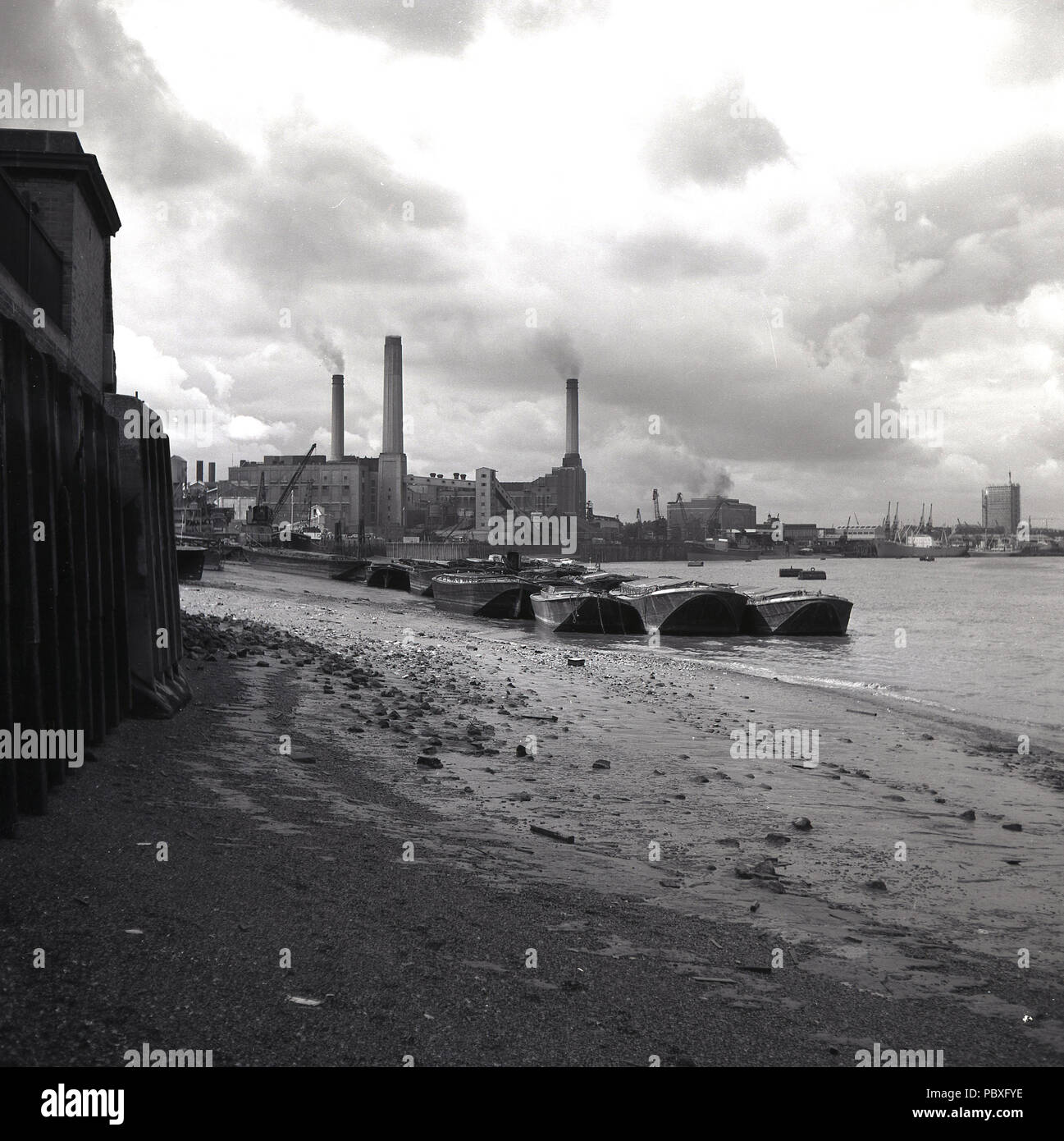 Degli anni Cinquanta, storico la marea è fuori come chiatte sedersi sulla riva sud del fiume Tamigi da una piccola spiaggia di ciottoli con una vista da est del lavoro di un Deptford Power Station in distanza, Londra, Inghilterra, Regno Unito. Progettato da Sebastian de Ferranti, Deptford è sapere come il primo al mondo il potere centrale stazione. Alimentate a carbone, era senza precedenti in entrambi la sua scala e alta tensione e ha continuato a funzionare fino alla fine degli anni sessanta. Foto Stock