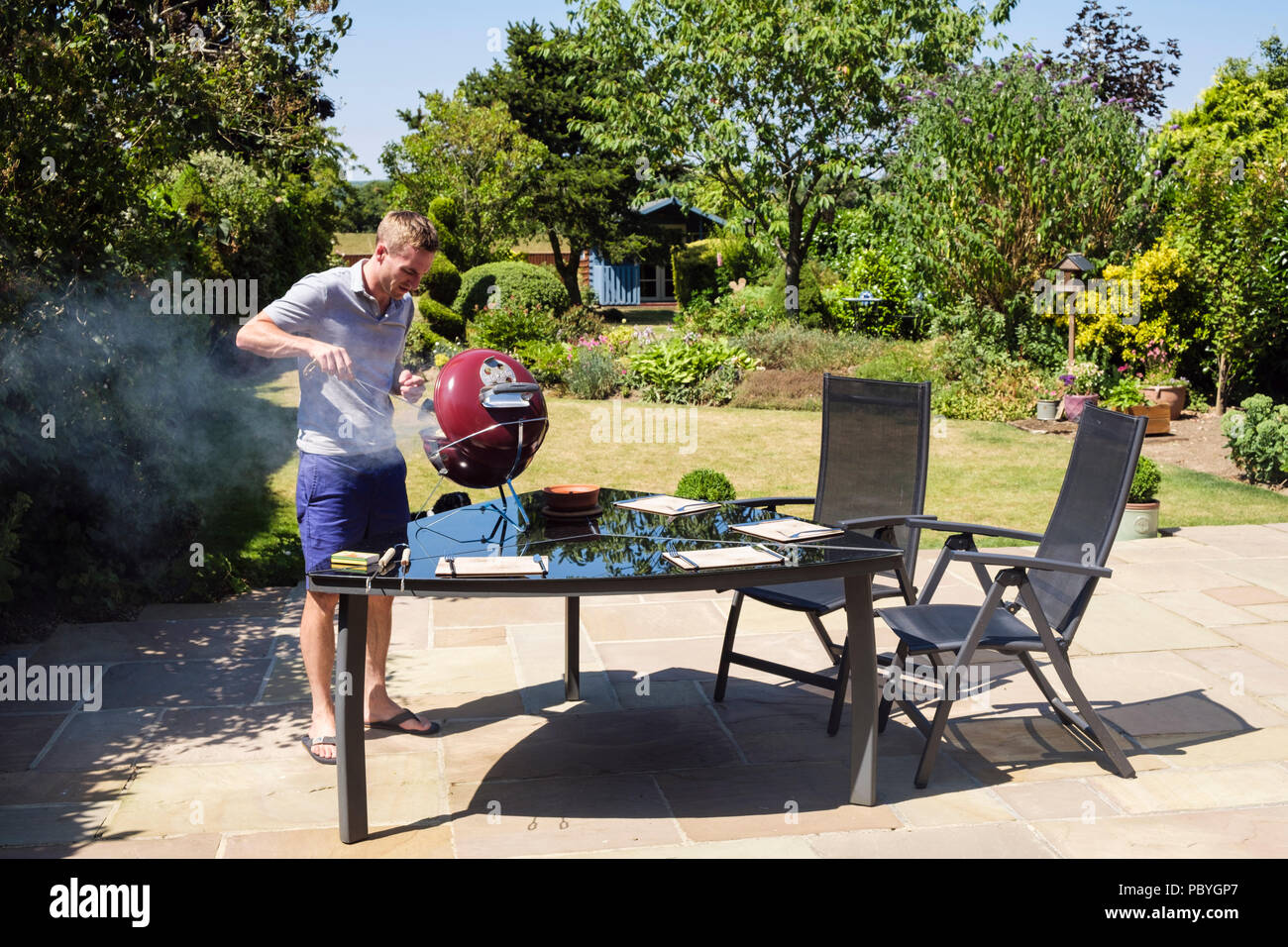 Autentica immagine di un giovane uomo Millenario che cucina su un barbecue su un patio giardino cortile tavolo in una calda stagione estiva del 2018. Inghilterra, Regno Unito, Gran Bretagna Foto Stock