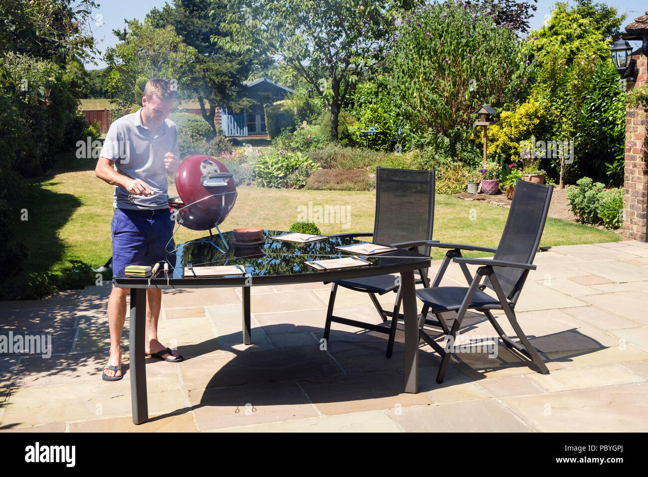 Autentica immagine di un giovane uomo Millenario cuoce su un barbecue su un tavolo giardino cortile interno su un patio in calda estate del 2018. Inghilterra, Regno Unito, Gran Bretagna Foto Stock