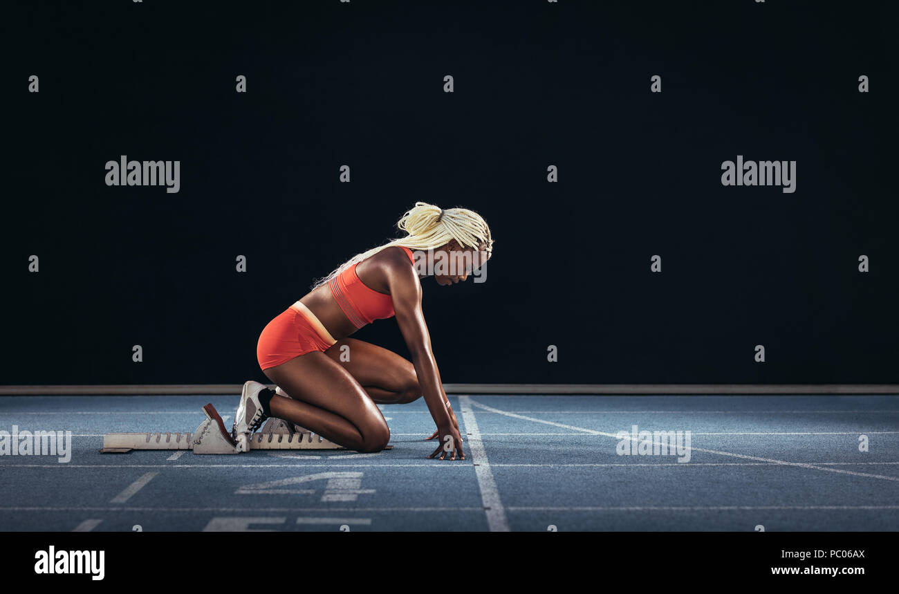 Atleta femminile di prendere posizione sul suo segna per iniziare la corsa. Vista laterale del femminile ottenere pronto all'inizio linea sulla via di corsa su un bl Foto Stock