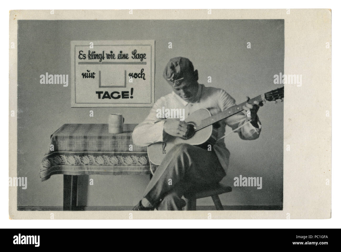 German Historical photo Cartolina: soldato nel tappo di foraggio suona la chitarra seduto su una sedia. L'esercito di svago, seconda guerra mondiale, la Germania, il Terzo Reich Foto Stock