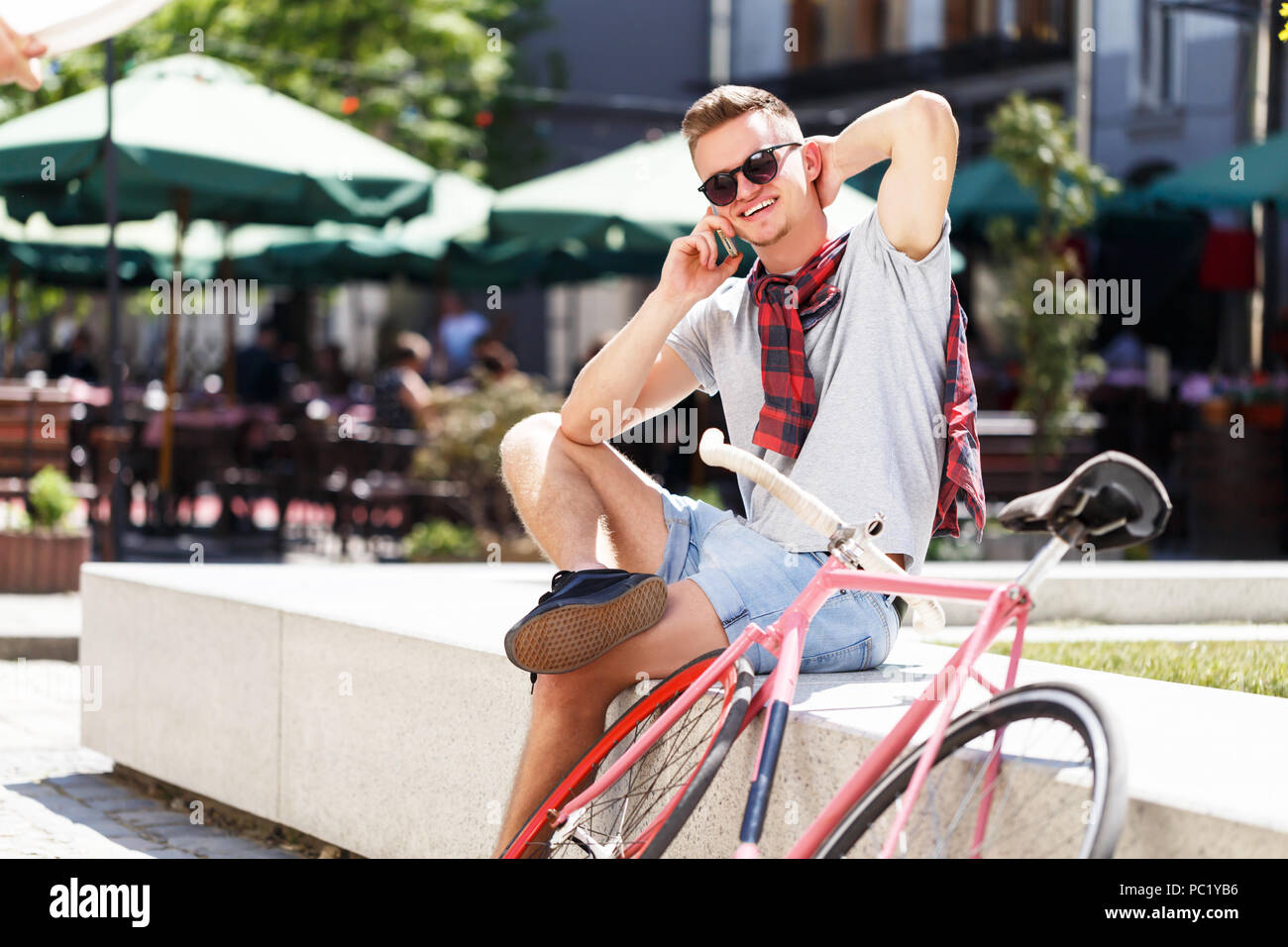 Bel giovane hipster uomo in occhiali da sole seduta vicino alla bicicletta e utilizza lo smartphone nel soleggiato street Foto Stock