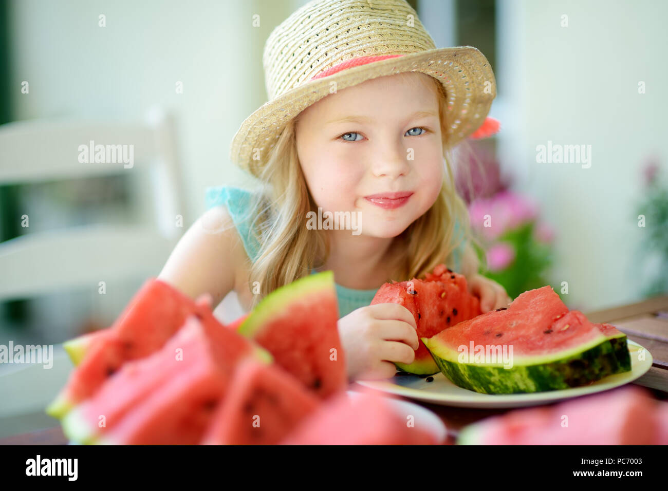 Funny bambina indossa cappello di paglia di mordere una fetta di cocomero all'aperto sul sole e caldo giorno d'estate. Un sano cibo organico per bambini piccoli. Foto Stock