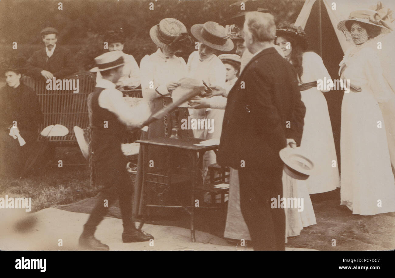 Vintage fotografia di un misterioso Edwardian riunione sociale. Signora Lettura di una carta, Bell su una tabella e un ragazzo tenendo un bat o lungo attrezzo. Foto Stock