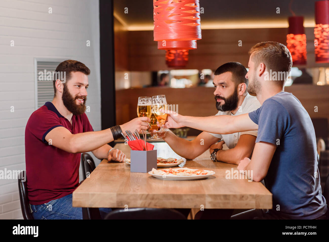 Gruppo di amici maschi tintinnio di bicchieri di birra gustosa prima di bere. Uomo Barbuto avente gustosa pizza sul loro tavolo. Tre amici divertendosi e appendere fuori a pizzeria. Foto Stock