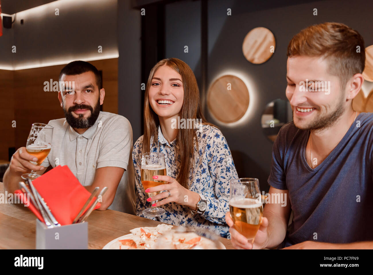 Vista frontale del barbuto due uomini e una donna sorridente, seduto e tenendo i bicchieri con la birra in mano. Amici divertendosi in bar o ristorante. Essi hanno anche mangiare pizza deliziosa. Foto Stock