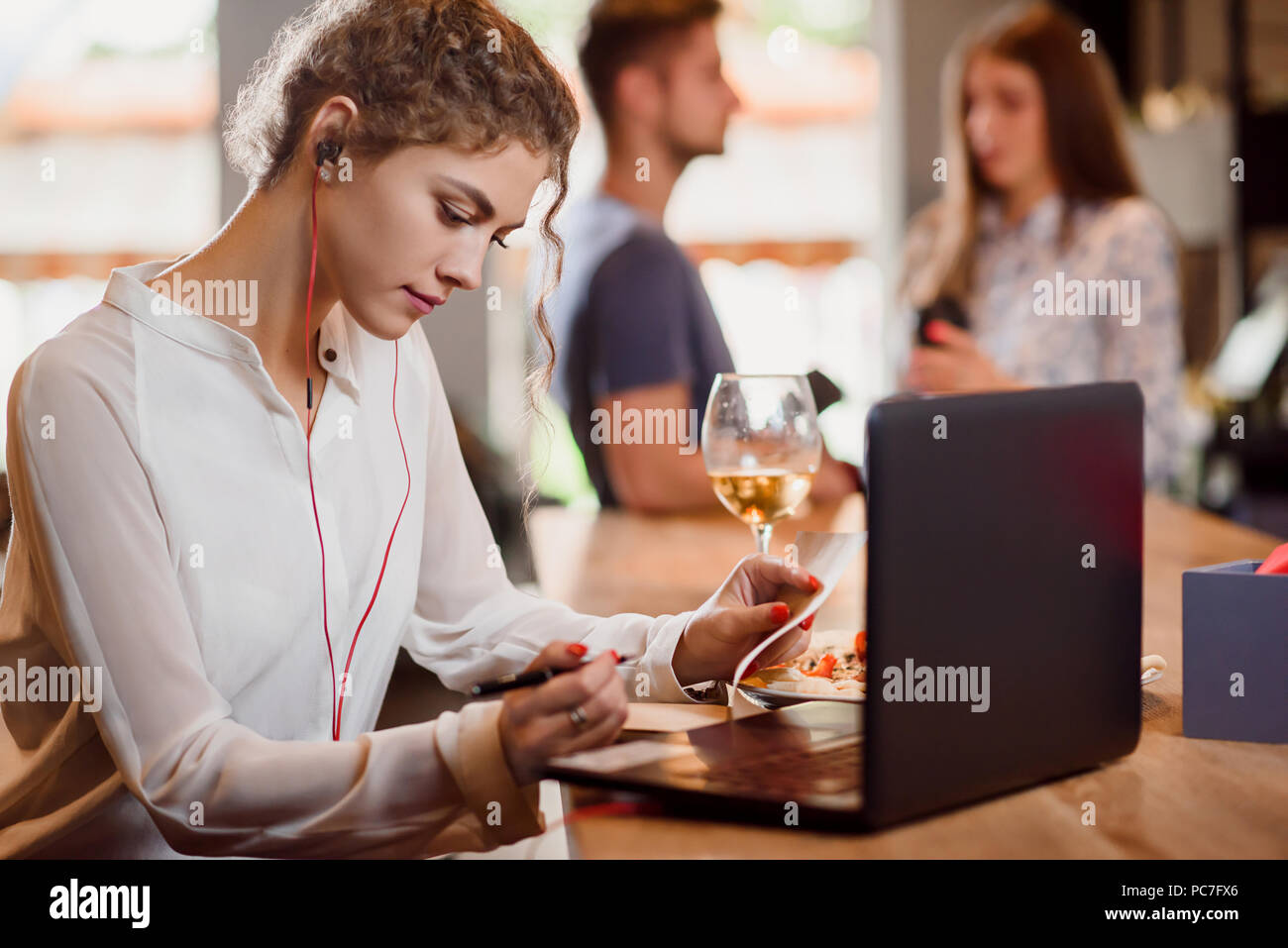 Stupendo giovane donna con la pelle liscia in seduta cafe e lavorare con il notebook. Donna con la pelle liscia e capelli ricci che indossa gli auricolari. e tenendo pen persone che parlano in background. Foto Stock