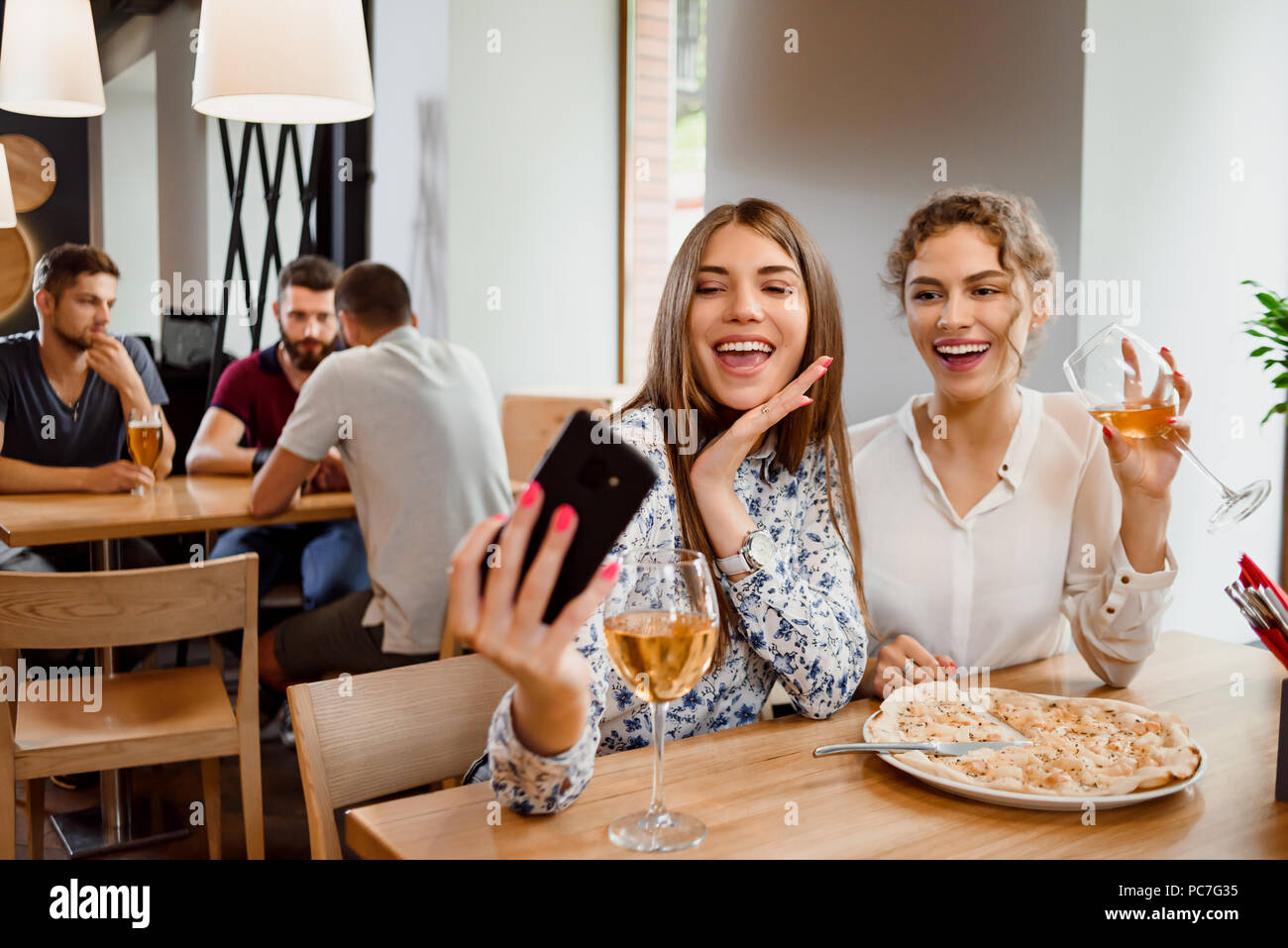 Due migliori amici facendo selfie, guardando il telefono e sorridente. Bella giovani donne per divertirsi insieme. Ragazze positiva di bere il vino bianco e avente una deliziosa pizza sul tavolo. Foto Stock