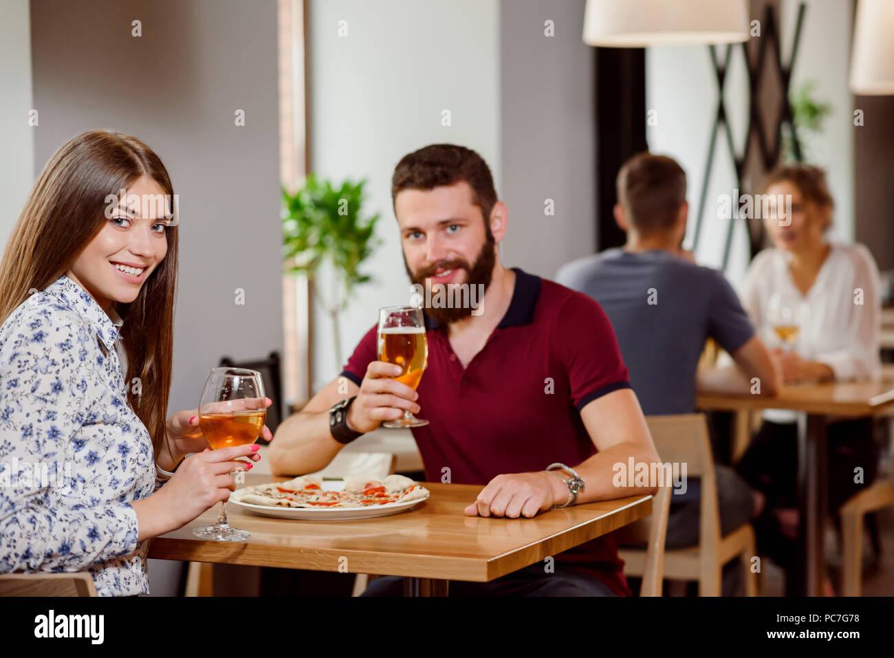 Bella giovane seduto in pizzeria e posa con bicchieri di vino bianco e birra. Splendida donna e uomo barbuto guardando la fotocamera e sorridente. Persone che parlano in ristorante sul background. Foto Stock