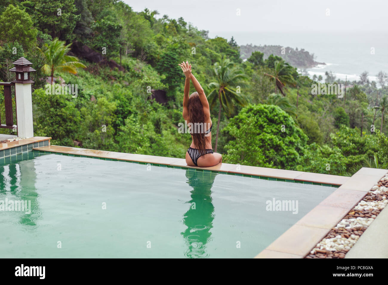 Vista posteriore di una ragazza con i capelli lunghi che pongono a piscina su resort tropicale Foto Stock
