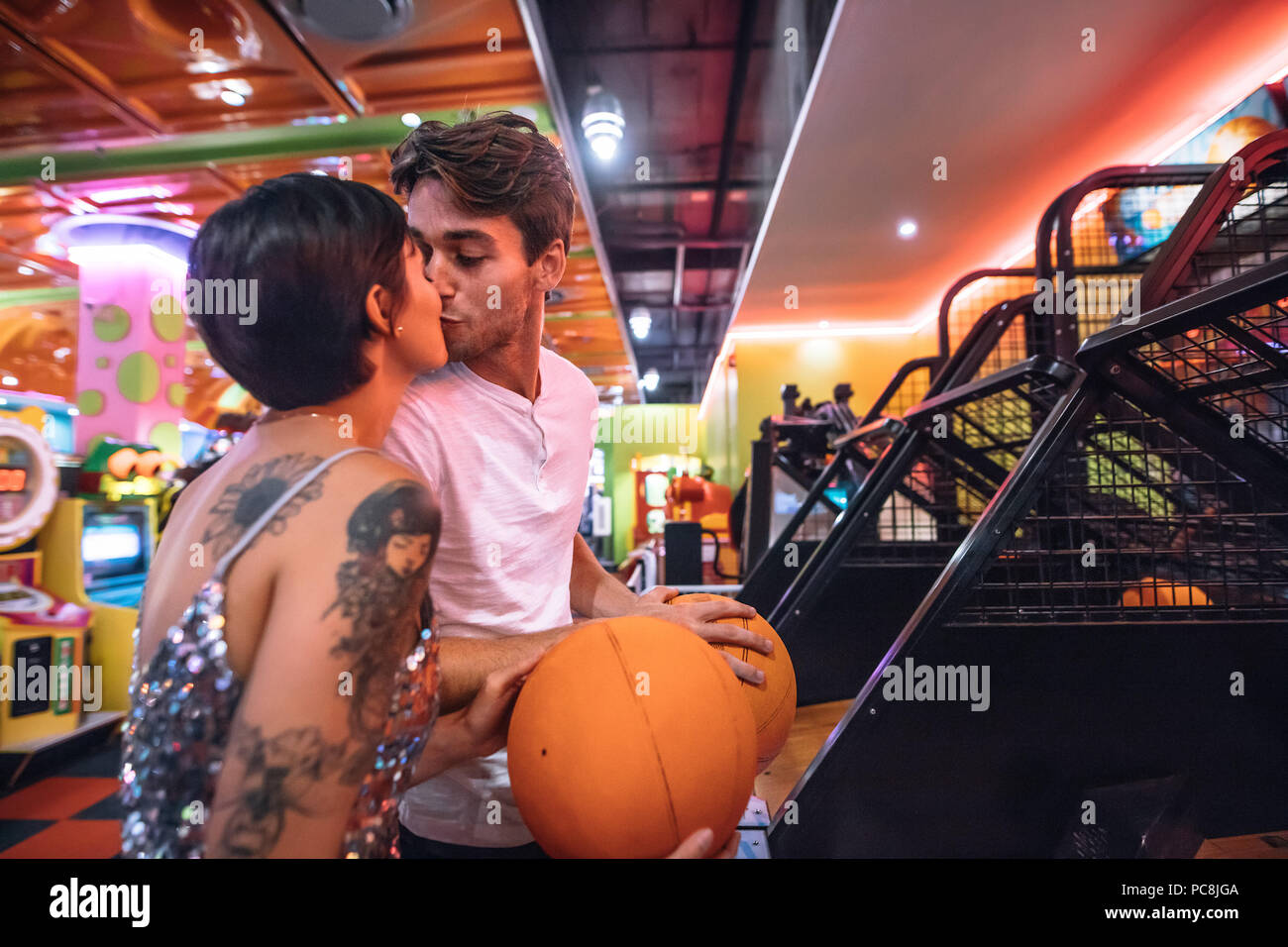 Paio di baciare ogni altro in piedi in un salotto di gioco tenendo basketballs. L uomo e la donna nella romantica atmosfera di un gioco arcade divertirsi giocando i giochi Foto Stock
