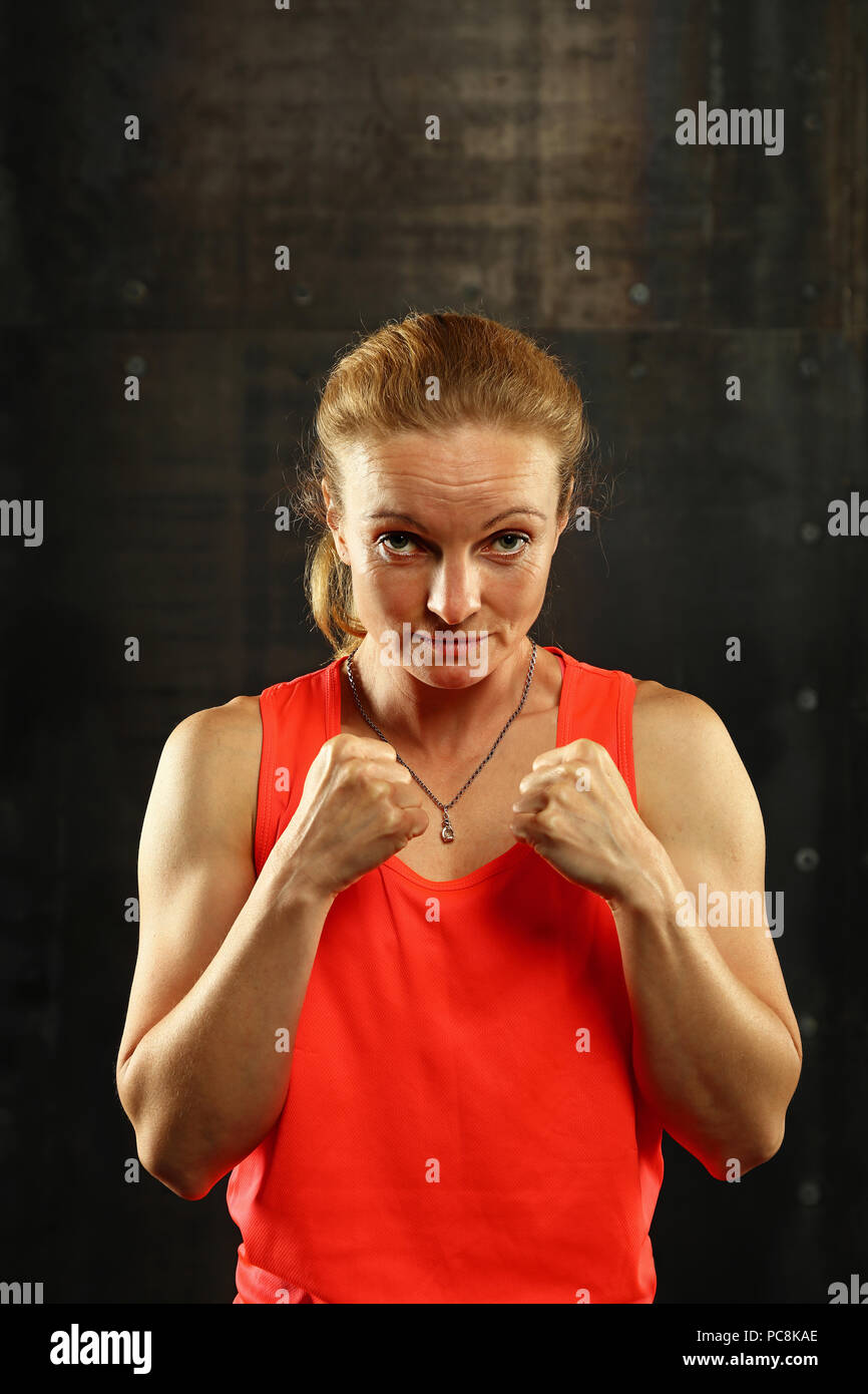 Close up front ritratto di una giovane età media athletic donna in abbigliamento sportivo in palestra su sfondo scuro, in piedi in atteggiamento di inscatolamento con mani e fis Foto Stock