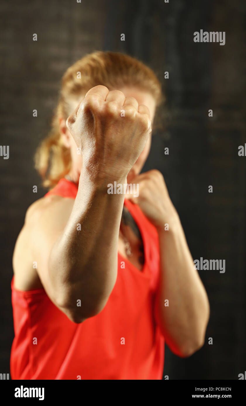 Close up front ritratto di una giovane età media athletic donna in abbigliamento sportivo in palestra su sfondo scuro, in piedi in atteggiamento di inscatolamento con mani e fis Foto Stock