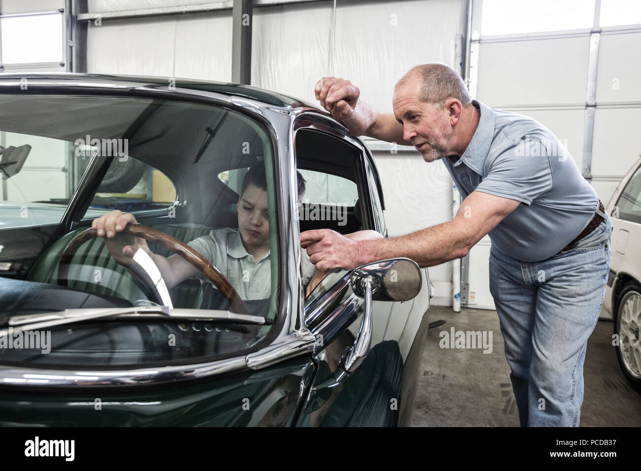 Un caucasico maschio senior meccanico di automobili mostra il suo nipote la vista del conducente dal sedile di guida di una vettura vecchia in un auto classica officina di riparazione. Foto Stock