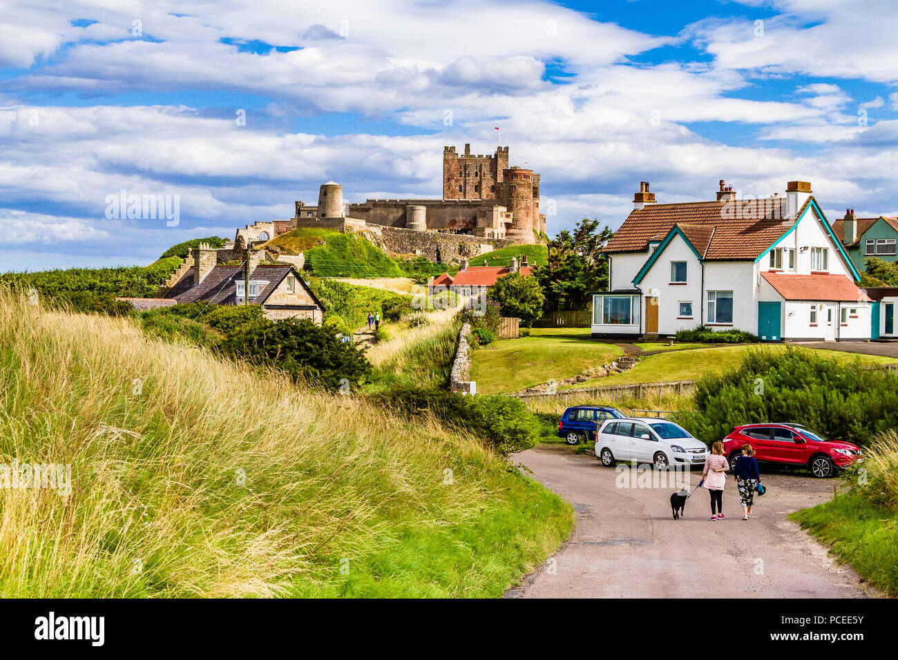 Due donne cane a piedi dalla spiaggia di Bamburgh down road a Bamburgh village, con il castello di Bamburgh in background. Bamburgh, Northumberland, Regno Unito. Foto Stock