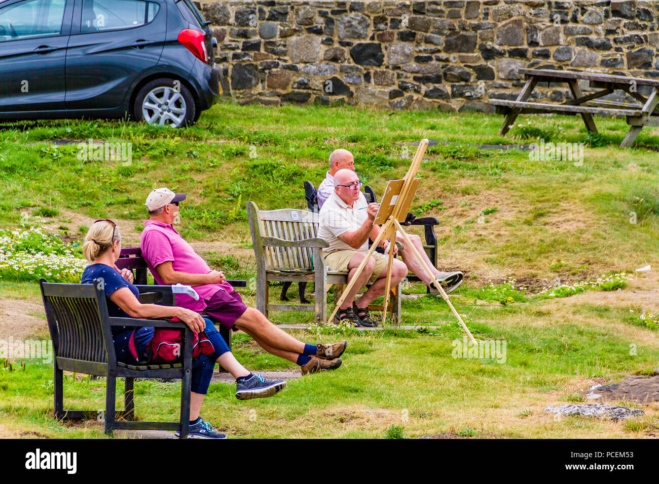 Le persone a rilassarsi sulle panchine nel nord-est del villaggio Craster guardando un uomo anziano vernice durante la canicola di agosto 2018. Craster, Northumberland. Foto Stock