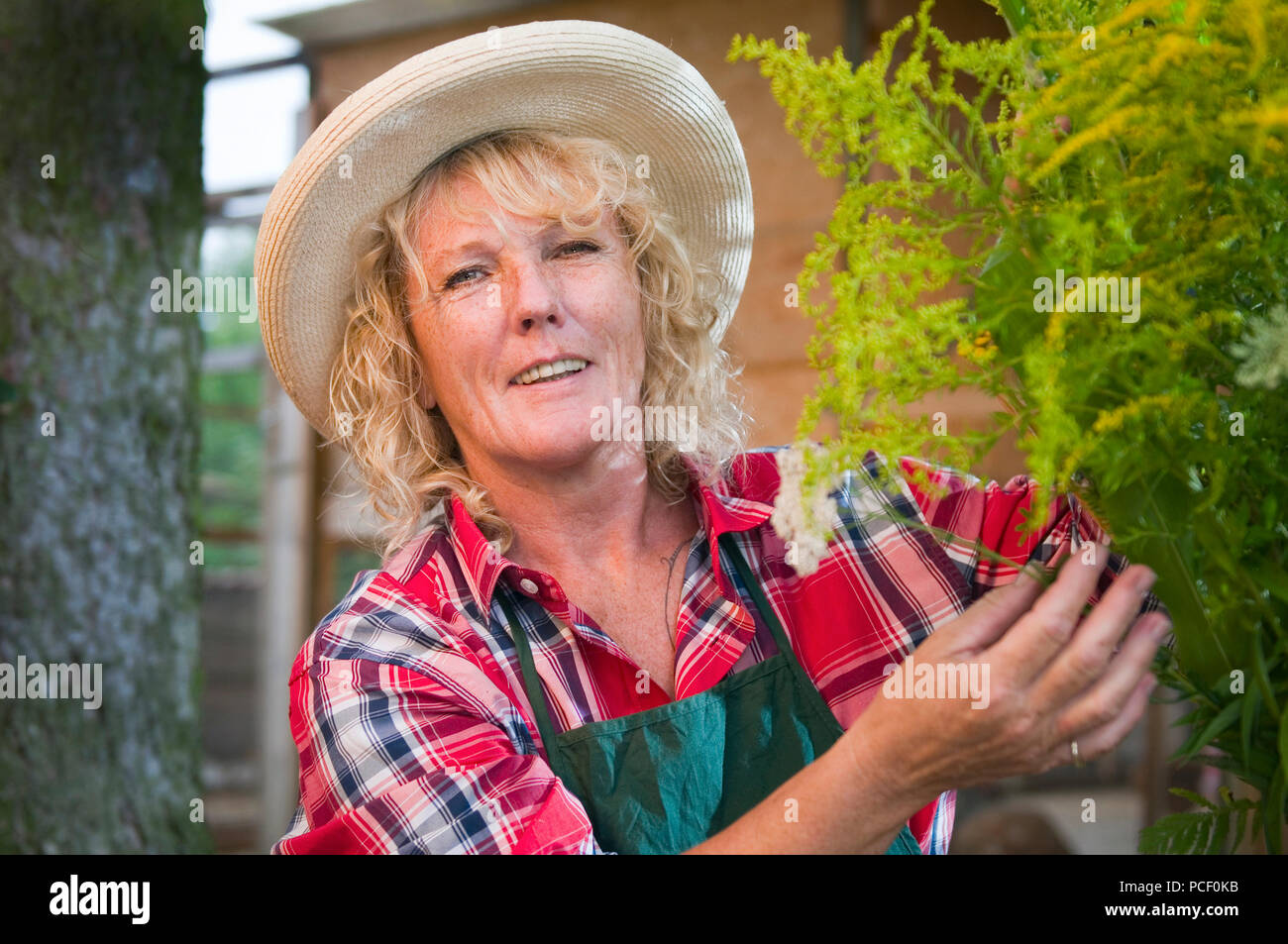 Testa-e-spalla ritratto di una donna bionda con le lentiggini e cappello di paglia in plaid shirt toccando un mazzo di fiori nel giardino e sorridente felicemente. Foto Stock