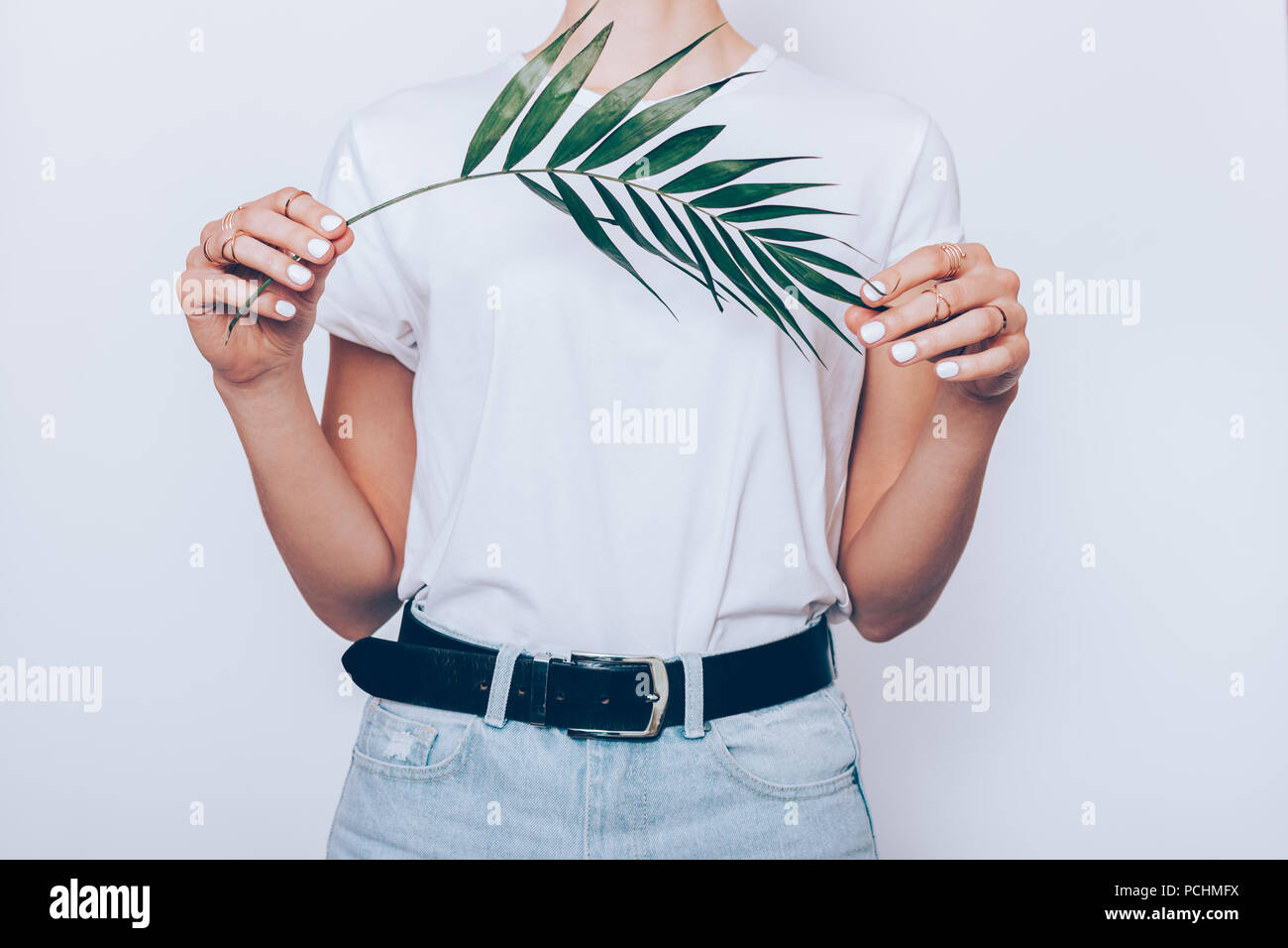 Snello giovane donna indossa blue high waist jeans con la cinghia e la pianura di t-shirt in piedi su sfondo bianco azienda verde foglia di palma, close-up. Foto Stock