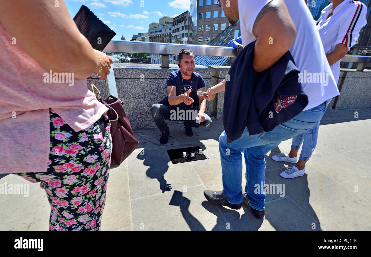 Uomo esecuzione illegalmente le tazze e i trick a sfera / tre gioco shell, sul London Bridge, Londra, Inghilterra, Regno Unito. La maggior parte dei "folla" sono membri del suo tè Foto Stock