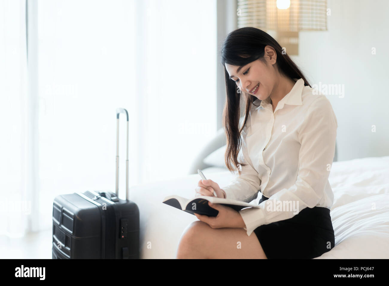 Close up di un felice bella Asian business donna sorridente allegramente in appoggio al suo hotel suite durante il viaggio di affari rendendo note e pensando strate Foto Stock